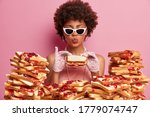 Small photo of Funny hungry woman eats tasty bread sandwich, keeps lips rounded, dressed elegantly, wears trendy sunglasses, enjoys tasty lunch, has good appetite and gluttony, comes closely to pile of toasts