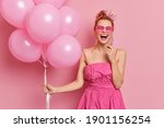 Cheerful redhead woman dressed in pink dress and sunglasses has fun on graduation party poses with bunch of inflated balloons laughs out gladully poses indoor. Female spends free time on prom night