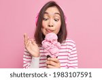 Asian brunette young woman looks at appetizing ice cream licks lips eats yummy frozen dessert listens music via headphones dressed in striped jumper isolated over pink background. Mmm how tasty
