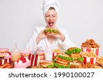 Small photo of Cheat mea and gluttony. Funny young woman exclaims loudly keeps mouth wide opened eats tasty burger surrounded by variety of fast food wears bathrobe towel over head isolated over white background