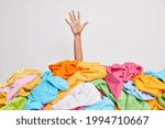 Unrecognizable human raises arm reaches out heap of colorful unfolded clothes busy doing wardrobe cleaning isolated over white background. Woman buried under cluttered clothing items. Decluttering