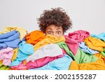 Small photo of Shocked woman with curly Afro hair stares bugged eyes drowned in huge pile of colorful clothing cleans out closet selects clothes for donation or recycling white background. Housework concept