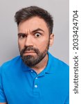 Small photo of Close up portrait of serious bearded adult man looks attentively at camera has scrupulous expression wears casual blue t shirt poses against grey background listens something very attentively