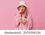 Small photo of Photo of positive young female model reads funny story or anecdote in internet holds modern mobile phone touches nose laughs happily wears hat and jumper poses against pink studio background