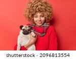 Good looking cheerful woman with blonde curly hair wears casual jumper and scarf around neck poses with dog going to have walk with favorite pet isolated over red background. People animals concept
