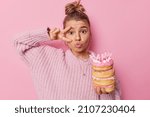 Lovely woman with hair gathered in bun keeps lips rounded makes peace gesture over eye holds birthday doughnuts celebrates special occasion wears knitted sweater isolated over pink background