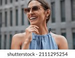 Happy young brunette European woman holds chin looks away smiles broadly being in good mood wears trendy sunglasses and fashionable clothes poses against blurred background spends free time in city