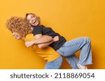Small photo of Happy optimistic women foolish around give each other piggyback ride laugh and have fun dressed in casual t shirts and jeans isolated over yellow background. Friendship relationship concept.