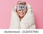 Small photo of Unrecognizable cheerful woman wears sleepmask on eyes smiles broadly wrapped in duvet expresses positive emotions being in good mood isolated over pink background. People rest and bedtime concept