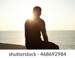 Silhouette of pensive African young male sitting on sidewalks at seaside watching sun setting over sea after outdoor training, waiting for his girlfriend, thinking about his relationships. Flare sun