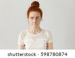 Indoor shot of cute redhead girl looking away, having doubtful and indecisive face expression, pursuing her lips as if forbidden to say anything. Confused young female posing isolated at white wall
