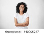 Small photo of Charming young dark-skinned woman with curly hairstyle having shy smile posing in studio in closed posture, keeping arms folded, feeling constrained and a bit nervous. Human emotions and feelings