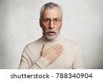 Small photo of Frightened shocked bearded man has beard stares at camera with bated breath, hears shocking news, can`t believe in them, isolated over white concrete background. People, pension, reaction concept