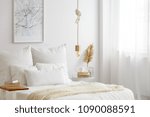Simple bulb lamp on a rope hanging above bed with white bedclothes, books and gold fern leaf on an end table in white bedroom interior