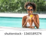 Portrait of happy fashion woman with sunglasses standing in swimming pool drinking cocktail. Beautiful african glamour girl with fresh soft drink for appetizer in luxury pool looking at camera.