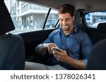 Happy smiling business man typing message on phone while sitting in a taxi. Young businessman in formal clothing using smartphone while sitting on back seat in car. Cheerful guy messaging with cell.