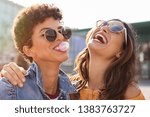 Small photo of Young latin woman laughing while friend inflating bubble gum. Closeup face of multiethnic friends enjoying outdoor street. Brazilian girl laughing and blowing chewing gum with friend embracing her.