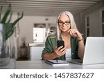 Happy senior woman using mobile phone while working at home with laptop. Smiling cool old woman wearing eyeglasses messaging with smartphone. Beautiful stylish elderly lady browsing site on cellphone.