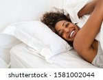 Cheerful young woman lying on bed playing with blanket and looking at camera. African girl feeling fresh after nap on bed with copy space. Laughing woman having fun while embracing pillow and blanket.