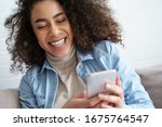 Cheerful afro american millennial woman holds smart phone watching social media stories video. Happy young mixed race teen girl using funny mobile app laughing, playing game, chatting on cell at home.