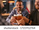 Happy mid adult friends clinking with beer mugs in pub. Three cheerful guys drinking draft beer, celebrating meeting and smiling. Laughing young men enjoying cold pint of beer during night at bar. 