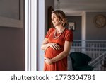 Small photo of Portrait of mature pregnant woman at home looking outside the window during maternity. Thoughtful pregnant woman thinking about the future family. Smiling lady in gestation caressing her baby bump.