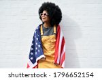 Happy young African American woman wrapped in USA flag laughing standing against white wall. Smiling free mixed race gen z hipster girl with Afro hair celebrating freedom on Independence Day concept.
