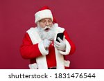 Surprised funny amazed old bearded Santa Claus wearing costume holding cell phone using mobile app on smartphone shocked by Christmas promotion, xmas applications ads isolated on red background.