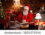Portrait of happy old kind bearded Santa Claus wearing hat, glasses, looking at camera, working on Christmas eve sitting at cozy home table late with presents, tree and candles preparing for holidays.