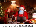 Old bearded Santa Claus wearing face mask, holding gift box on xmas eve sitting at cozy home table late in night using laptop computer. Merry Christmas Covid 19 coronavirus social distance concept.