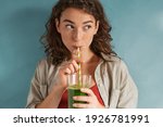 Beautiful woman drinking an organic green smoothie. Fit young woman drinking detox juice using paper straw isolated against blue background. Healthy girl enjoy detox drink and looking away. 