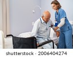 Small photo of Lovely nurse assisting senior man to get up from bed at hospital. Caring nurse supporting elderly patient while getting up from bed and move towards wheelchair at nursing home.