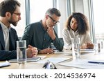 Focused doubtful mature businessman reading contract document thinking considering risks with professional lawyers legal experts executive team analyzing financial report sitting at office table.