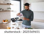 Young man preparing healthy breakfast indoors at home, pouring milk shake into glass.