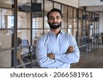 Confident happy successful ceo financial manager. Bearded indian businessman looking at camera standing in modern office with arms crossed. Handsome classy corporation owner. Business portrait.