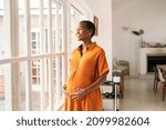 Mid black pregnant woman standing near window at home and thinking about her future family. Smiling african american lady with hands on belly imagine the growth of his baby. Middle aged pregnant woman