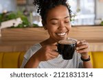 Small photo of Smiling black young woman smelling freshly brewed coffee with eyes closed in cafeteria. Beautiful african girl smiling while relaxing in a coffee shop. Close up face of girl drinking latte coffee.