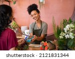 Smiling florist holding card reader machine at counter with customer paying with credit card. Young african american florist shop assistant holding payment machine while buyer purchase a bunch flower.