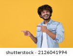 Small photo of Happy excited young indian man looking at camera pointing aside with fingers hand gesture at copy space advertising new promotion, presenting sale offer standing isolated on yellow background.
