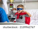 Small photo of Cheerful strong little boy wearing blue eyeband and red cape like superhero playing with nurse. Playful child gesturing dressed in superhero costume at clinic overcome adversity and health challenge.