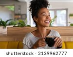 Carefree african american woman sitting in cafeteria drinking coffee while looking away. Black young woman drinking tea while thinking. Smiling girl relaxing and thinking while drinking cappuccino.