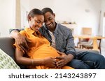 Small photo of Happy african american husband and pregnant woman hugging belly. Smiling black man hugging happy pregnant wife sitting on sofa and holding tummy. Mature loving couple expecting their first baby.
