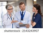 Small photo of Senior woman doctor having a discussion in hospital hallway with medical staff. Doctor discussing patient case status with his team. Pharmaceutical representative showing medical report on tablet.