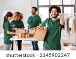 Small photo of charity, donation and volunteering concept - happy smiling male volunteer with food in box pointing to camera over international group of people at distribution or refugee assistance center
