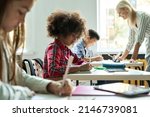 Small photo of Diligent concentrated hard working diverse miltiethnic classmates schoolchildren writing tasks in notebooks with happy teacher explaining and helping. Schoolwork in diversity team. Education concept.