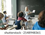 Small photo of Smiling female teacher showing new maths task to elementary middle diverse schoolchildren sitting on desk at chalkboard holding tablet device. Kids using laptops. Education tech concept.