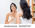 Woman caring of her beautiful skin on the face standing near mirror in the bathroom. Young woman applying moisturizer on her face. Smiling girl holding little jar of skin cream and applying lotion.