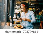 Young African woman smiling while sitting alone at a counter in a bistro taking photos of her meal with her smartphone