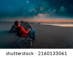 man sitting in a camping chair alone on the beach looking at the starry night next to a tent