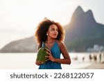 Small photo of waist up portrait cheerful young brazilian afro hairstyle woman walking on the beach holding a coconut water in Ipanema